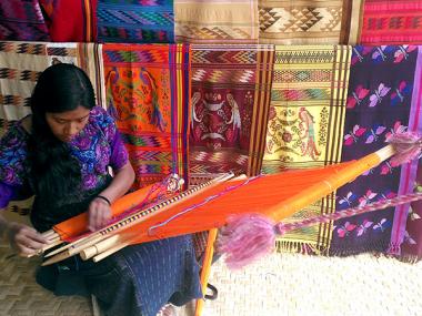 Woman using a traditional weave with many completed fabrics hanging in the background